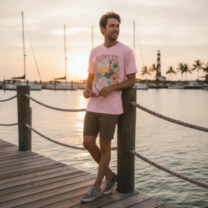 A man stands on a wooden dock at sunset wearing a light pink graphic T-shirt with 'JUPITER' and tropical artwork, olive shorts, and gray sneakers, with water, boats, and a lighthouse in the background.