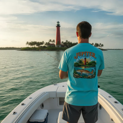 A man on a boat seen from behind, wearing a light blue t-shirt with a large graphic print on the back that reads 'JUPITER MARINE' and shows a dolphin and tropical scene. In the background is a red lighthouse on an island.