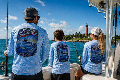 Three people on a boat wearing matching light blue long-sleeve shirts with a large 'America 250' fishing graphic on the back, with ocean water and a lighthouse in the background.
