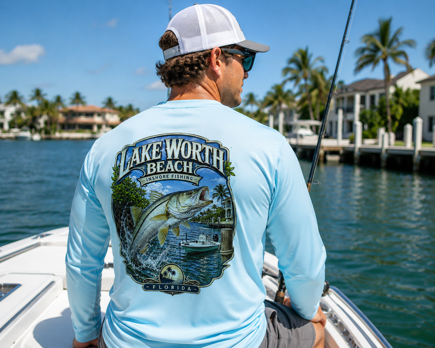 A man on a boat wearing a light blue long-sleeve performance shirt with a large Lake Worth Beach Florida graphic on the back, paired with a white cap and sunglasses, with water and waterfront houses in the background.