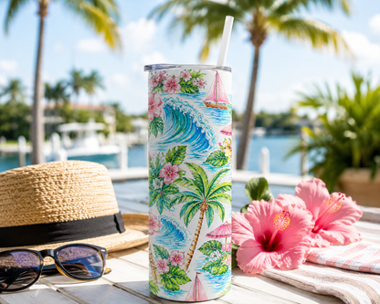 A tall insulated tumbler with a tropical wave and palm tree print stands on a table outdoors near a pool or marina. A straw hat, sunglasses, pink hibiscus flowers, and a striped towel are arranged around it, with palm trees and water blurred in the background.