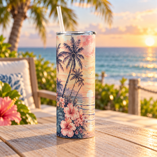 A tall insulated tumbler with a clear lid and straw stands on a wooden table outdoors at sunset, printed with palm trees and pink hibiscus flowers over an ocean scene. A blurred beach and lounge chair are in the background.