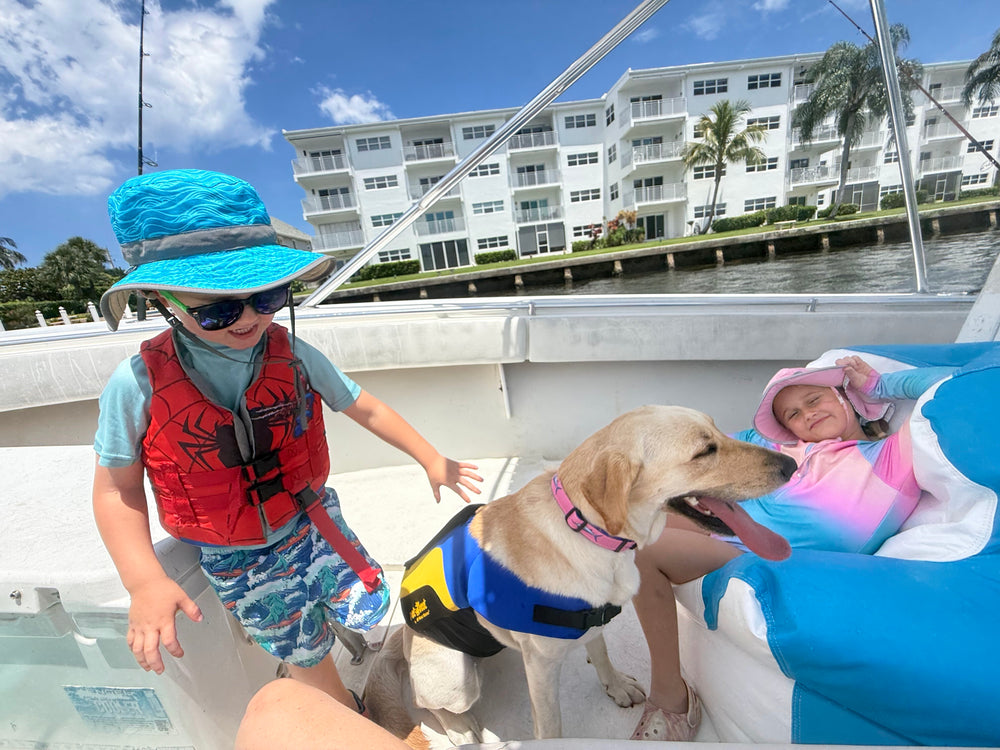 A child wearing a blue sun hat, sunglasses, and a red life vest stands on a boat next to a golden retriever dog wearing a blue and yellow life vest. Another child is lying on a blue and white float in the water behind them, with a white apartment building in the background under a blue sky.
