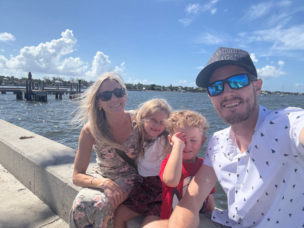 A family of four taking a sunny outdoor selfie on a waterfront promenade. The father on the right wears a white short-sleeve button-up shirt with a small black triangle pattern, a black cap, and blue mirrored sunglasses.