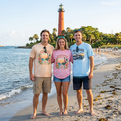 Three people standing on a sandy beach in front of a lighthouse, wearing graphic t-shirts. The person on the left wears a light peach t-shirt with a colorful graphic, the person in the middle wears a pink t-shirt with a graphic that says 'LOVE IT UP', and the person on the right wears a light blue t-shirt with 'PALM BEACH' printed on the front.