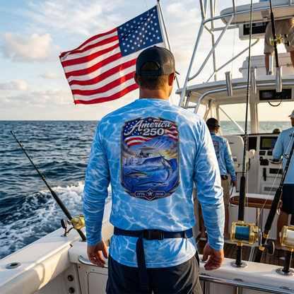 A man on a fishing boat seen from behind, wearing a light blue long-sleeve performance shirt with a large graphic on the back featuring a fish, a boat, and an American flag. The boat is sailing on the open ocean under a cloudy sky.