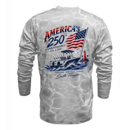 A long-sleeve performance shirt shown from the back, featuring a gray and white watercolor splash pattern. The back is decorated with a large graphic celebrating 'America's 250th Anniversary 1776 - 2026' with an American flag, a fishing boat, and a red lighthouse.