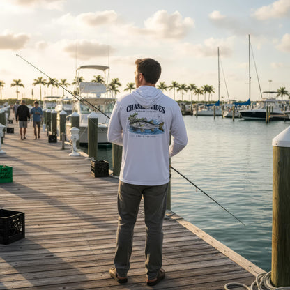 A man stands on a wooden dock at a marina, viewed from behind, wearing a white long-sleeve hoodie with a large fish graphic and the text 'CHASING ADES' on the back. He is holding a fishing rod, with boats, water, and palm trees in the background.