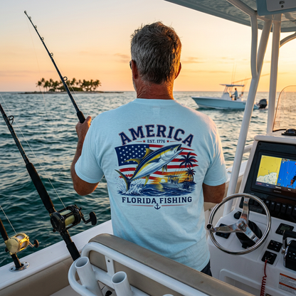 A man on a boat at sunset wearing a light blue t-shirt with a large graphic on the back that reads 'AMERICA FLORIDA FISHING' and shows a fish, American flag, and sunset scene.